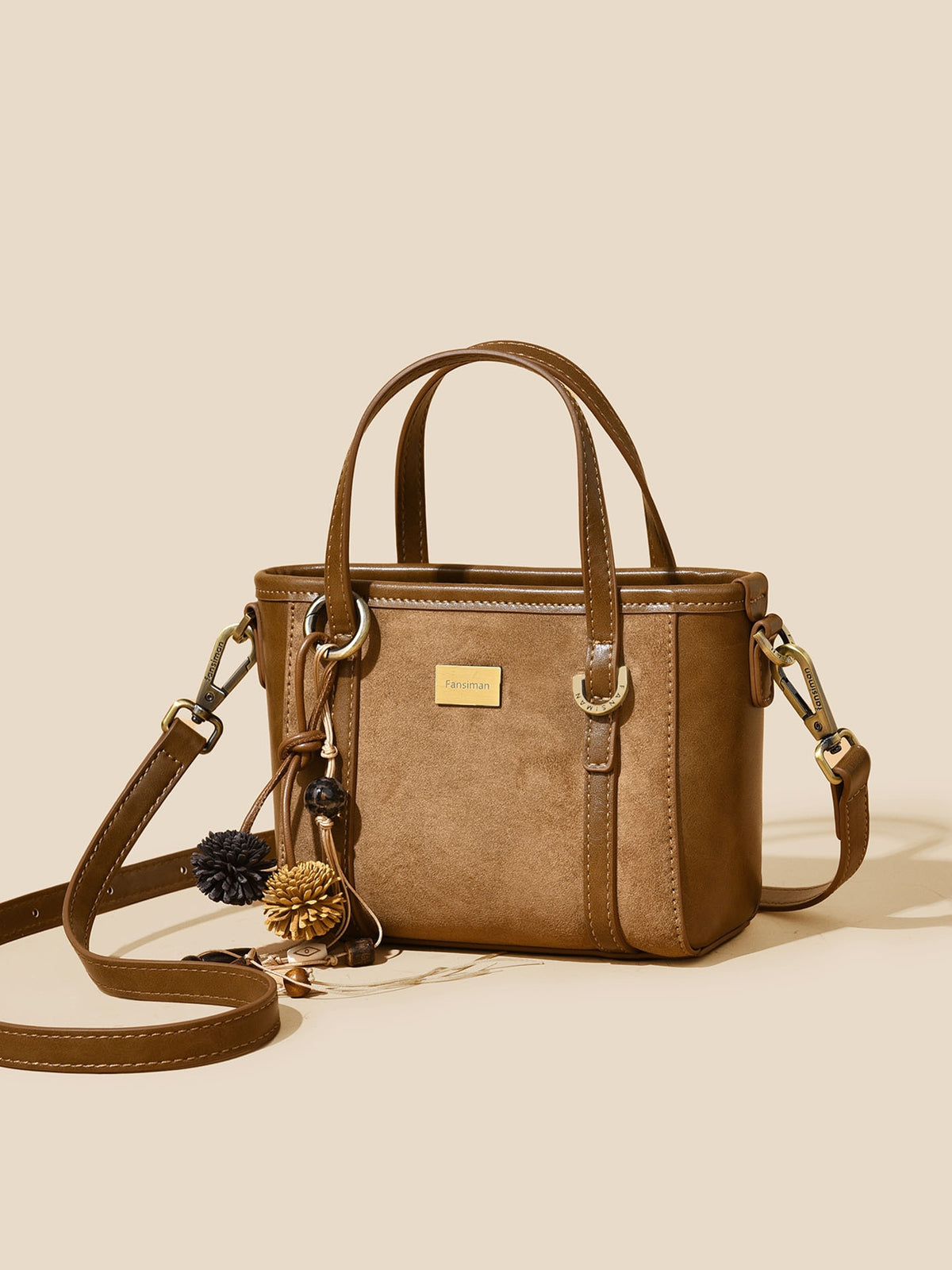A structured brown faux-leather handbag with dual top handles, a detachable shoulder strap, and decorative pom-pom and pinecone charms, displayed on a solid beige background.
