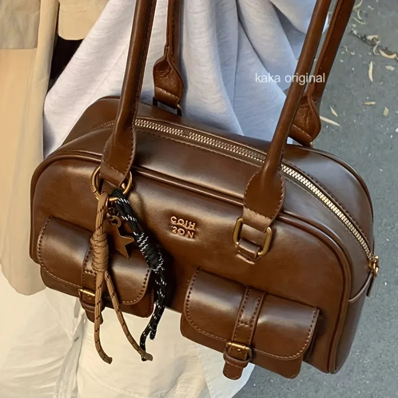 A brown faux-leather shoulder bag with a rounded rectangular shape, featuring dual top handles, a top zipper, front flap pockets, and a decorative braided tassel charm. The brand name 'COAH DON' is embossed on the front.