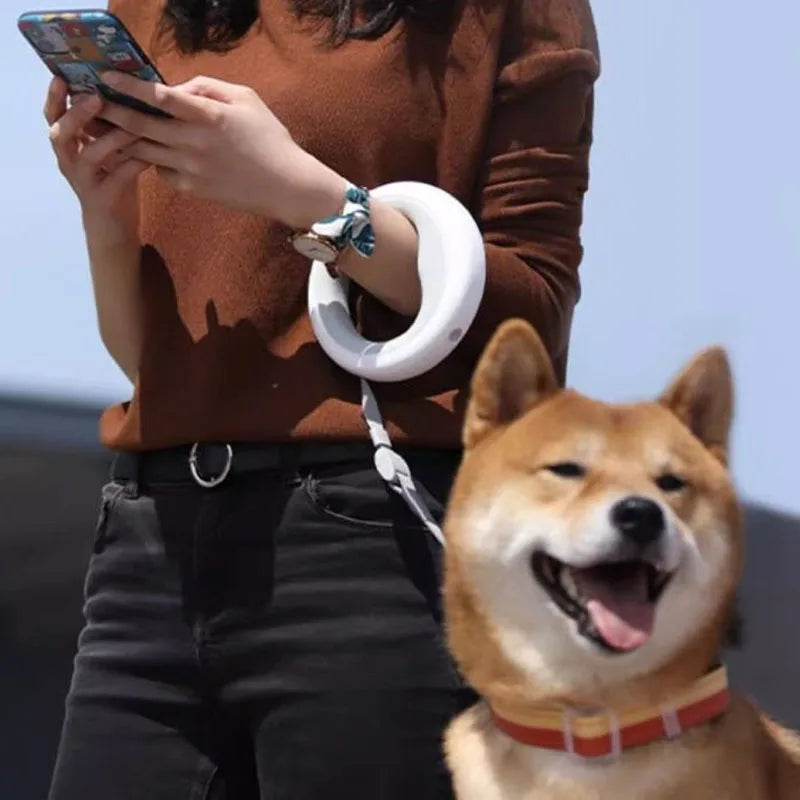 A Shiba Inu dog wearing a red and yellow collar attached to a white ring leash held by a person wearing a brown sweater and black pants, looking at their phone.