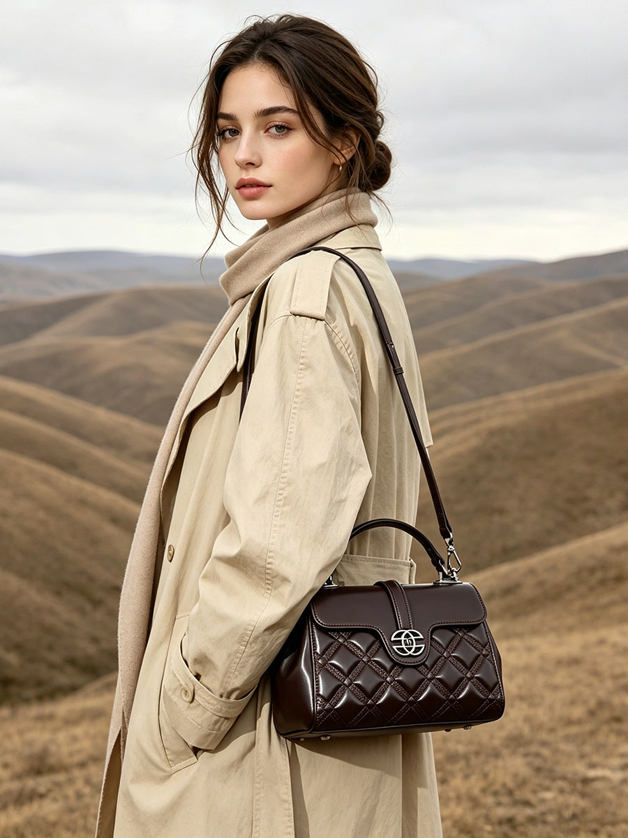A woman wearing a beige trench coat stands outdoors against rolling hills, carrying a dark brown quilted shoulder bag with a silver interlocking logo.