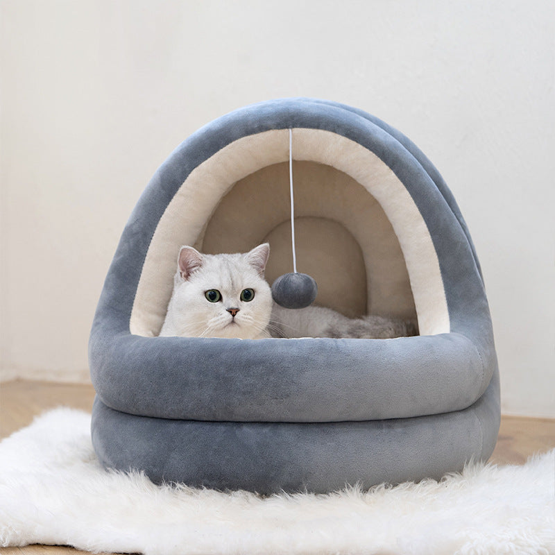 A white cat sits inside a plush dome-shaped pet bed in soft gray velvet with cream lining, placed on a white rug against a light wall.