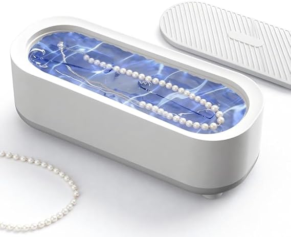 An oval-shaped white jewelry storage box with a transparent lid showing blue light inside, containing a pearl necklace. A second pearl necklace lies on the white surface beside it, and a matching white textured lid or cover is shown behind the main box.