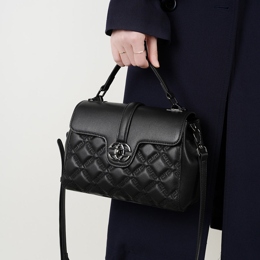 A black quilted leather handbag with a top handle and shoulder strap, held by a person wearing a dark navy coat against a light gray studio background.