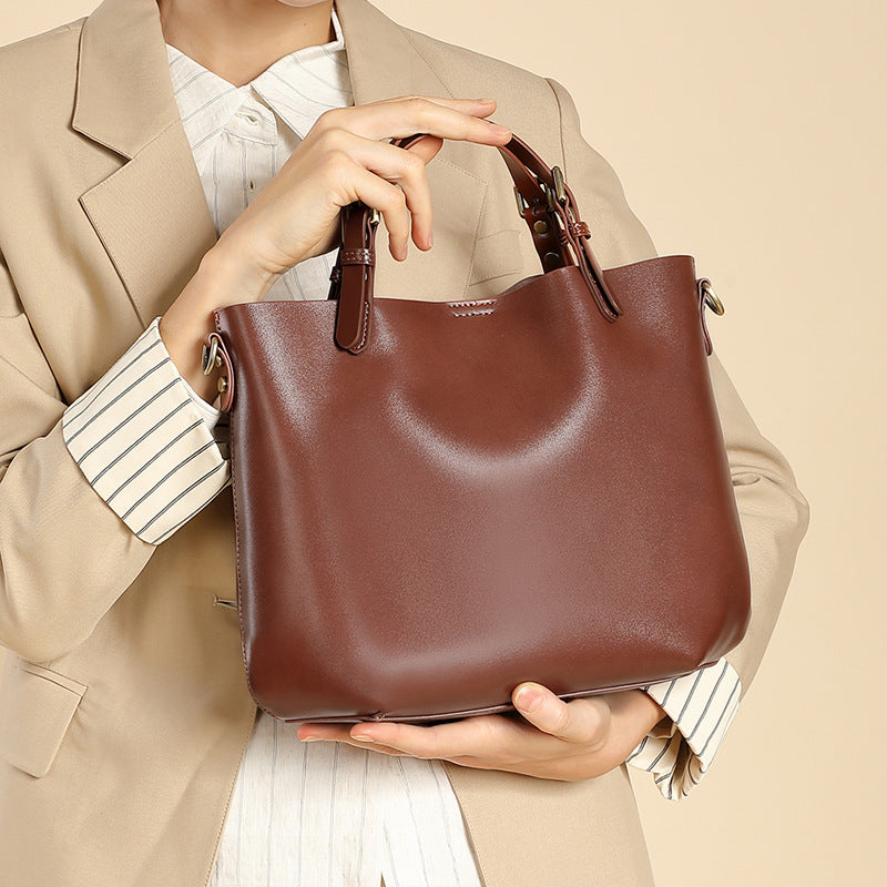 A woman in a beige blazer and striped shirt holds a brown leather tote bag with top handles against a plain beige background.