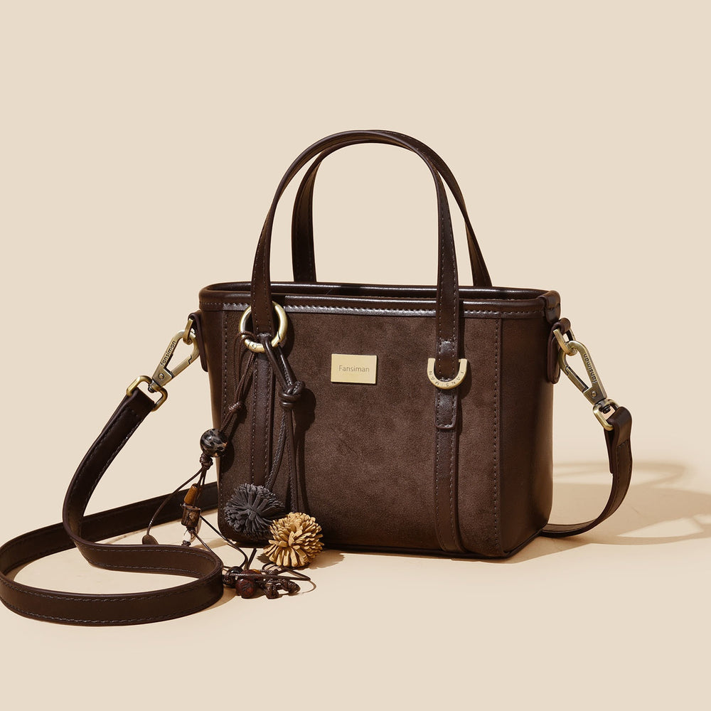 A dark brown leather tote bag with double top handles, a detachable shoulder strap, and decorative hanging charms, displayed on a warm beige background.