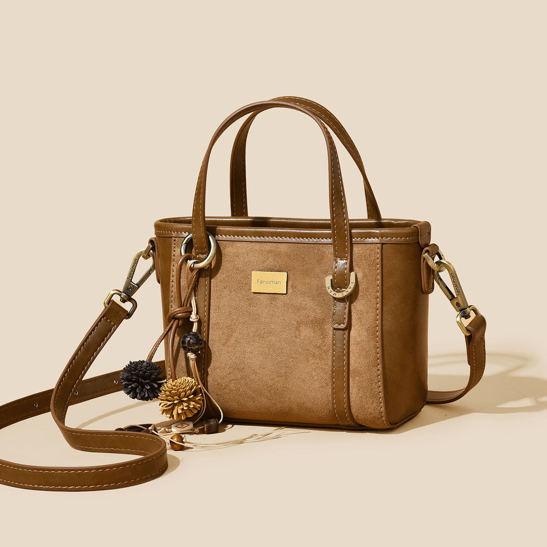 A structured brown faux-suede handbag with dual top handles, a detachable shoulder strap, and decorative pom-pom and pinecone charms, shown on a warm beige background.