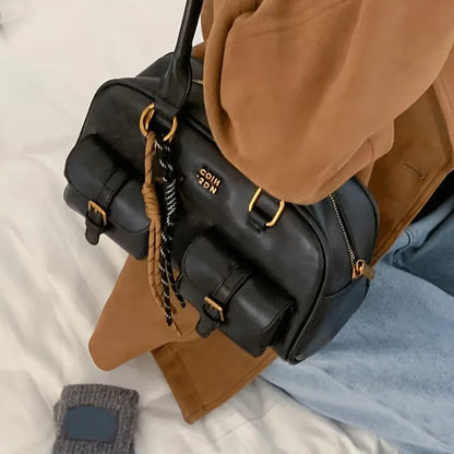 A black faux-leather shoulder bag with gold hardware and a braided keychain charm, styled on a white bed with a tan coat, light blue jeans, and a gray sock nearby.
