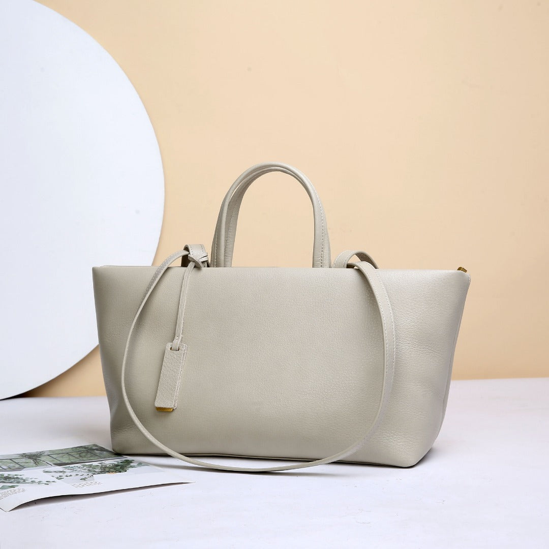 A light beige pebbled leather tote bag with dual top handles and a detachable shoulder strap, displayed on a white surface against a beige background with a white circular prop.