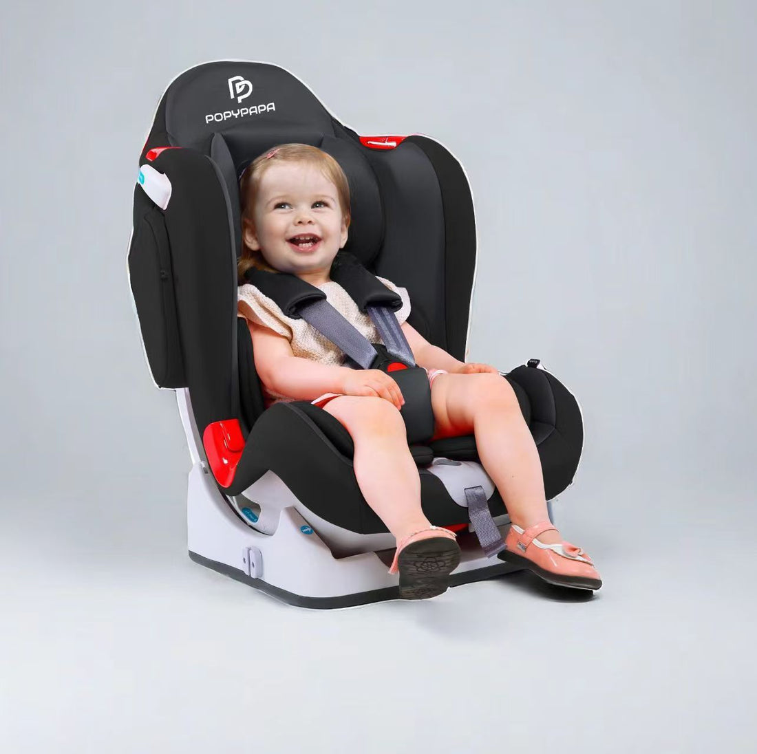 A smiling toddler is seated in a black and gray Popvava car seat with red accents, photographed on a clean light gray studio background.