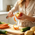 Person peeling an apple with a peeler on a wooden cutting board with vegetables.