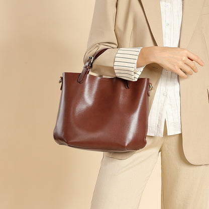 A person wearing a beige blazer and striped shirt carries a large brown leather tote bag against a plain beige background.