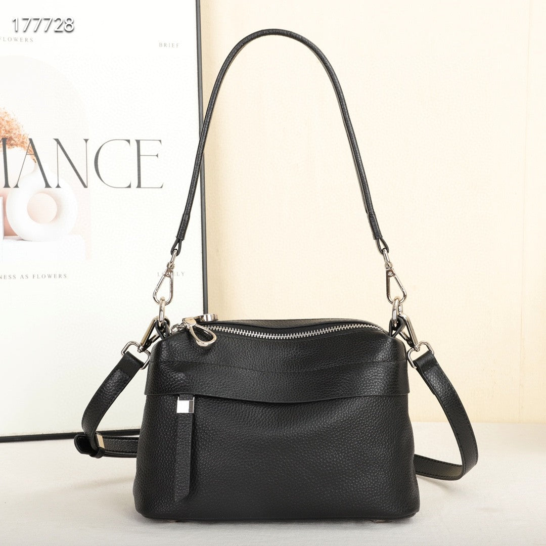 A black pebbled leather crossbody handbag with a top zipper, front flap pocket, and silver-tone hardware, shown against a light beige background with a magazine partially visible behind it.