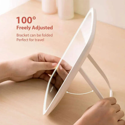 A white electronic makeup mirror with an adjustable stand is being held by two hands on a light wooden table. Red text on the left reads &