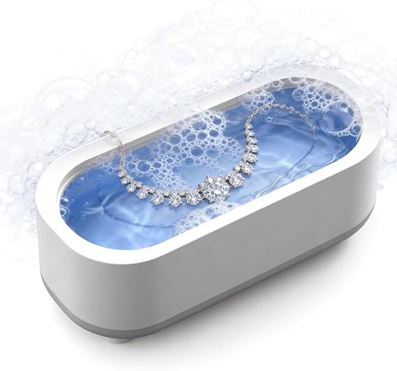 An oval ultrasonic jewelry cleaning bath with a gray outer shell and a clear top, filled with blue water and bubbles, holding a silver necklace with clear gemstones.