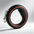 A black retractable dog leash with a bright orange accent ring and a silver metal clasp, shown on a light gray studio background. The brand name &