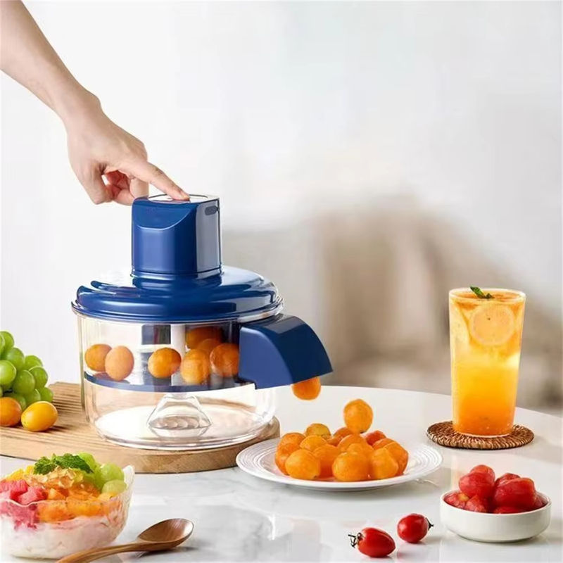 A hand is pressing the top of a blue and clear countertop food chopper, which is processing small orange fruits. The scene is set on a white table with a glass of orange juice, a plate of orange fruits, and other food items, against a soft, blurred background.