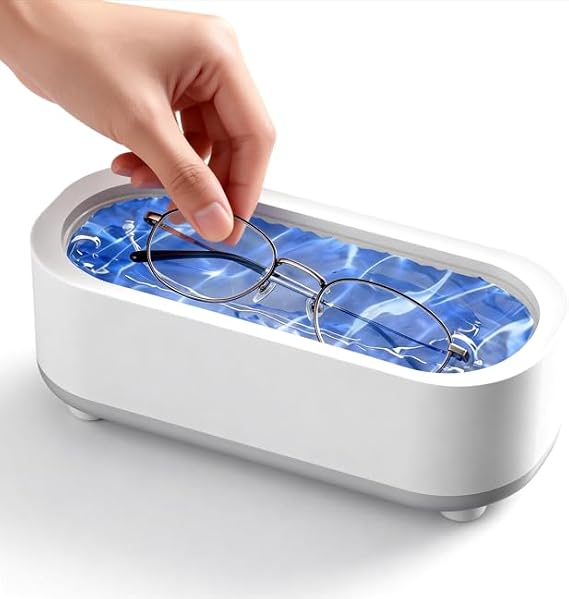 A hand placing a pair of eyeglasses into a white rectangular glasses cleaner tray filled with blue water and bubbles, on a clean white background.