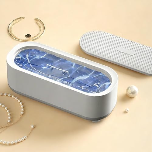 An oval-shaped jewelry organizer tray with a white exterior and a blue marble-patterned interior, shown with its matching lid on a beige background. The tray is styled with jewelry props including a gold crescent ring, pearl necklaces, and a pearl bead.