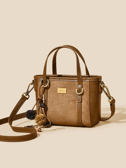 A structured brown faux-leather handbag with dual top handles, a detachable shoulder strap, and decorative pom-pom and pinecone charms, displayed on a solid beige background.