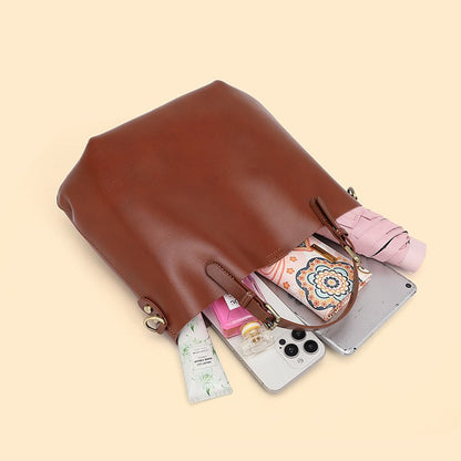A brown leather tote bag shown open with various items partially visible inside, including a patterned wallet, a pink compact, a tube of lotion, and a smartphone, on a solid pale beige background.