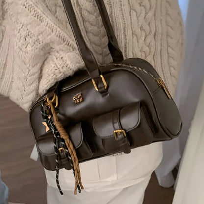 A dark brown leather shoulder bag with multiple front pockets, gold-tone hardware, and a braided charm, shown being worn over the shoulder by someone in a cream cable-knit sweater.