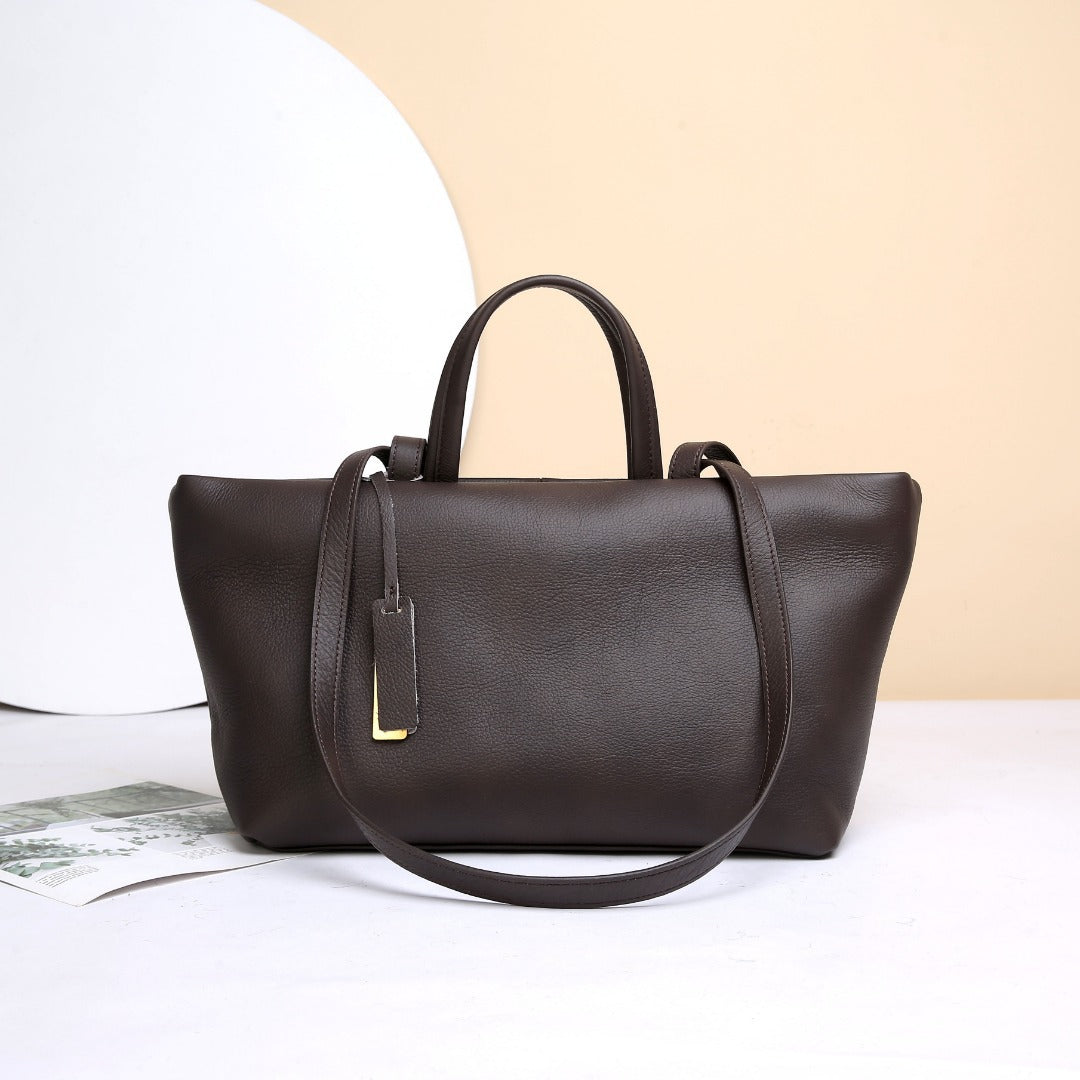A dark brown textured leather tote bag with double top handles and a long shoulder strap, displayed on a white surface against a beige and white background.